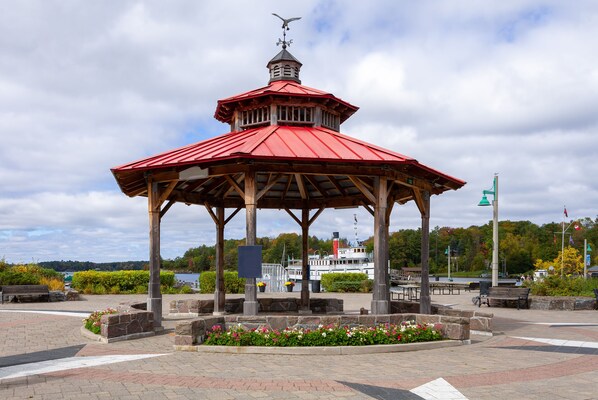A gazebo with a red roof and a weather vane, surrounded by stone planters and benches, with a river and boats in the background. - Residence Inn by Marriott Gravenhurst Muskoka Wharf (Gravenhurst)