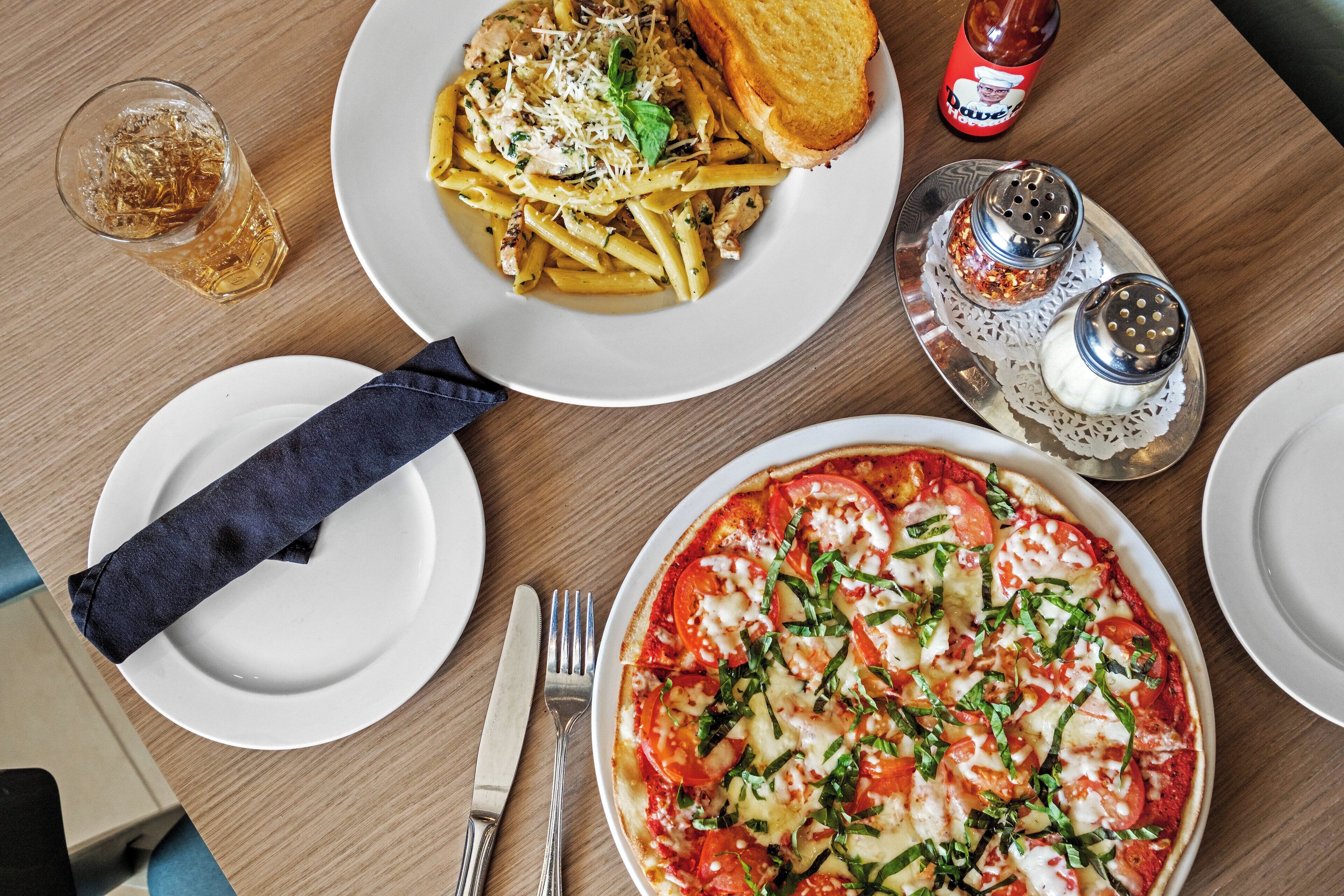 A dining table with plates of pasta and pizza, a glass of water, and condiments.