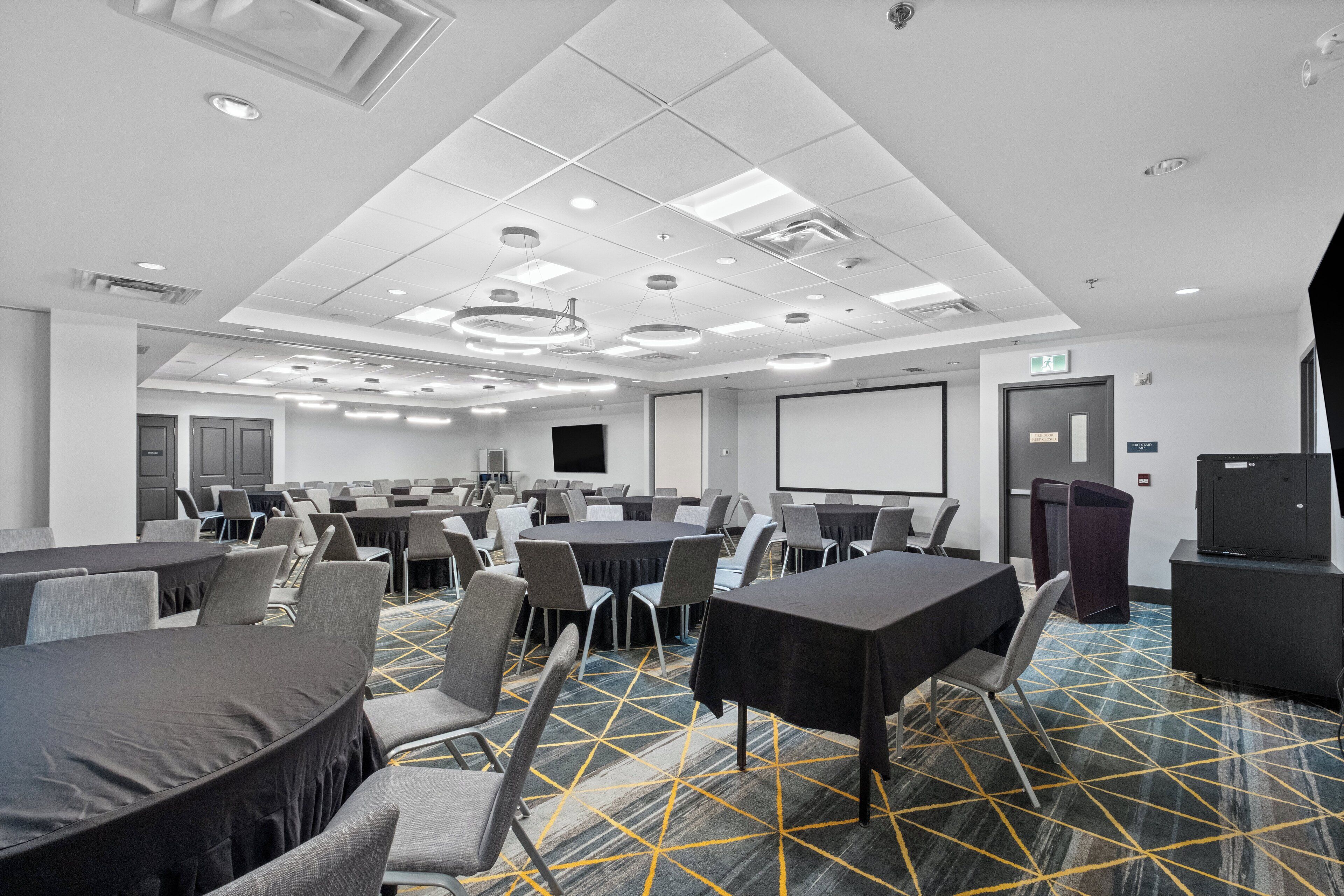 A conference room with multiple tables and chairs, a projector screen, and a presentation area.