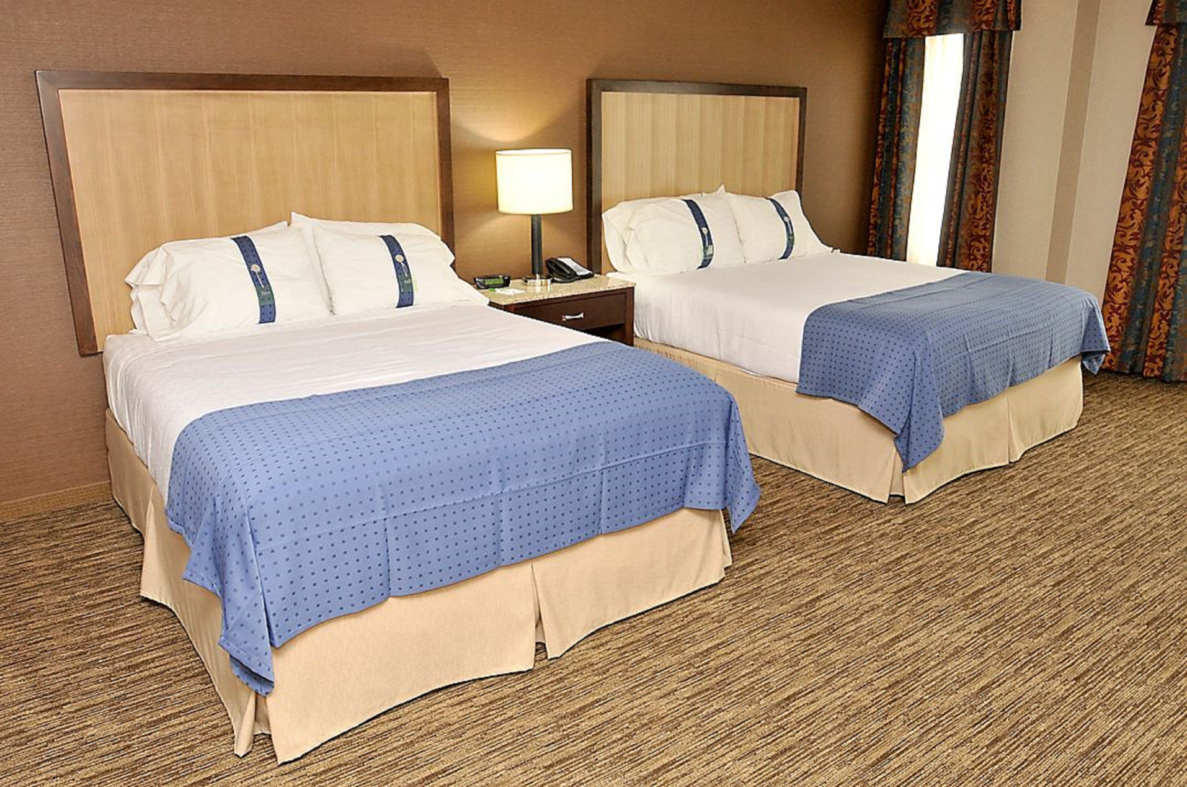 Habitación de hotel con dos camas, cada una con cubrecama azul y almohadas blancas. Entre las camas hay una mesita de noche con una lámpara y un teléfono.