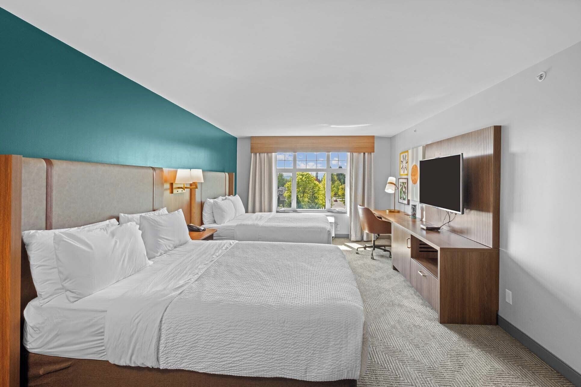 A hotel room with two beds, a desk, a television, and a window with a view of trees.