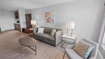 A modern living room with a grey sofa, a round coffee table, and a grey armchair. There is a painting on the wall, a small table with a lamp, and a kitchenette in the background.