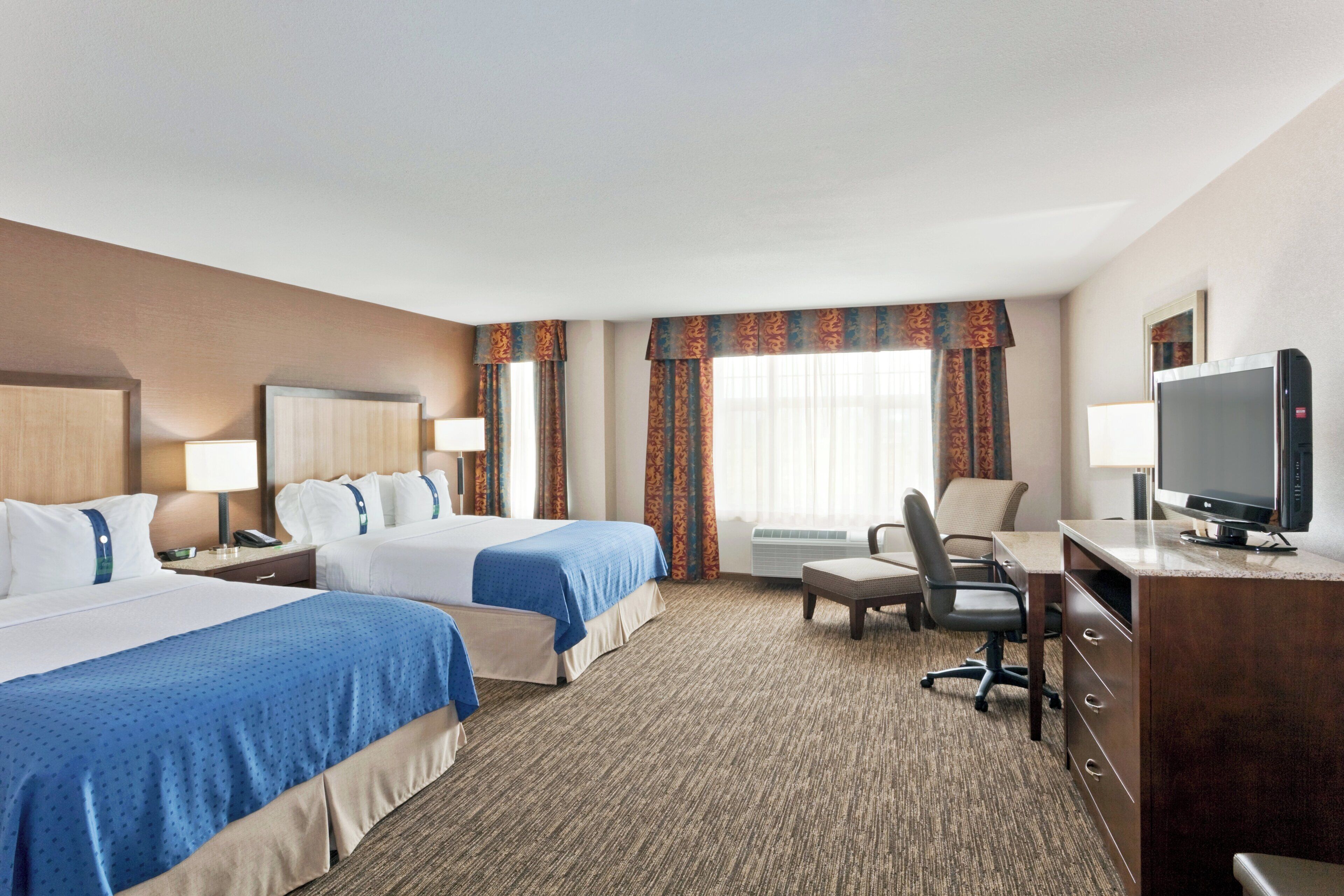 A hotel room with two beds, a desk, a chair, a television, and an air conditioning unit.