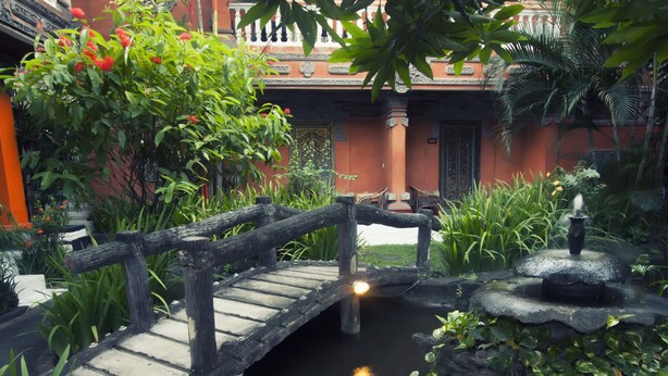 Un pont en bois enjambe un étang orné d’une fontaine, le tout étant entouré d’une végétation luxuriante et d’un bâtiment à l’architecture traditionnelle.