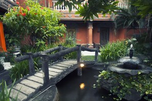 A wooden bridge over a pond with a fountain, surrounded by lush greenery and a building with traditional architecture. - Seminyak Paradiso Hotel (Seminyak)