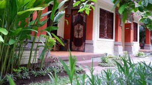 Il s’agit d’un bâtiment traditionnel à la façade rouge et blanche, doté d’une porte d’entrée décorative et d’un jardin agrémenté de diverses plantes.