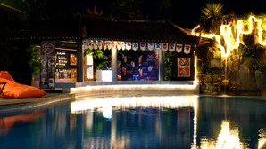 Un espace piscine nocturne agrémenté d’un bar de style tiki, illuminé de lumières chaleureuses, et d’une cascade.