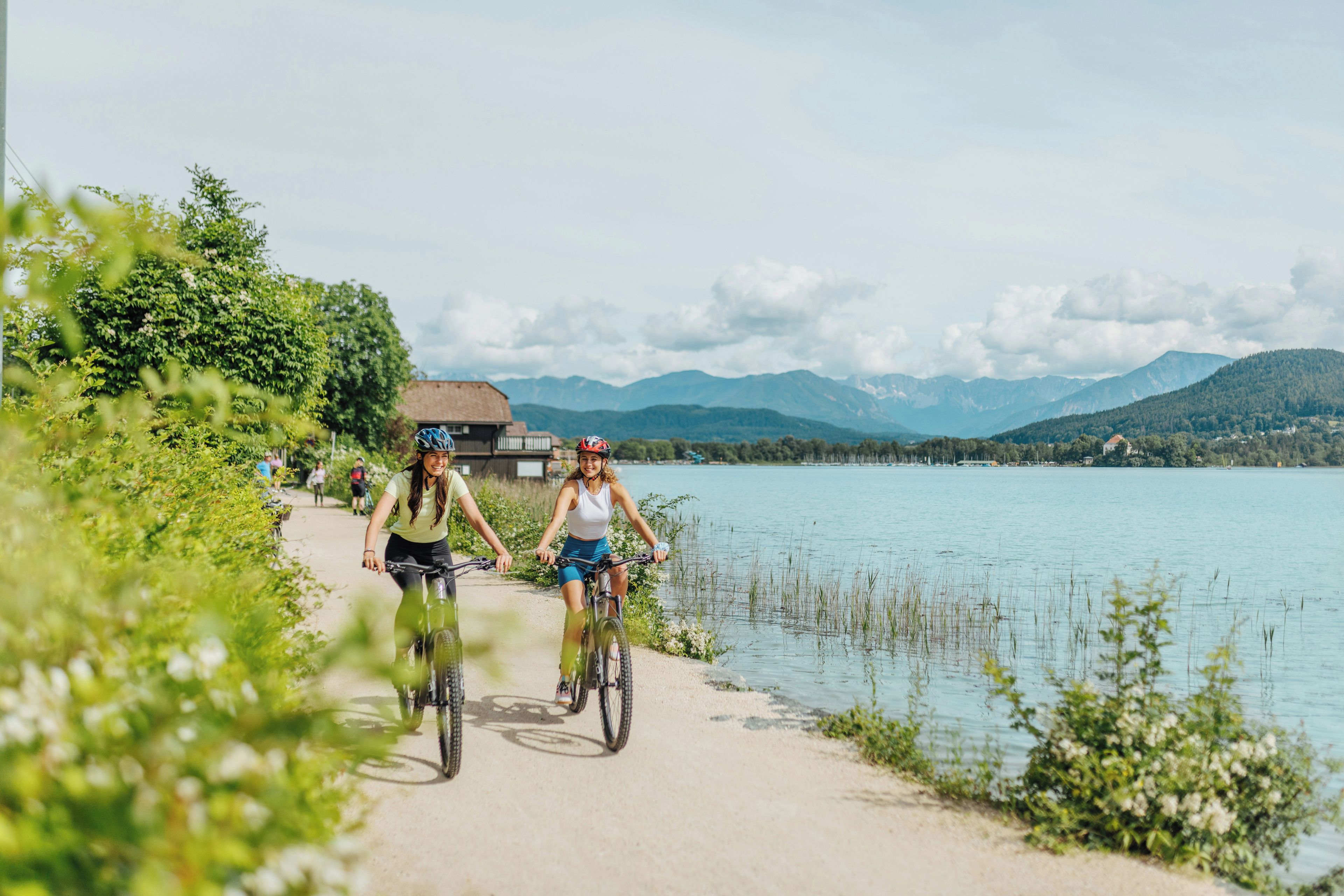 Dos personas andan en bicicleta por un sendero junto a un lago, con montañas al fondo.