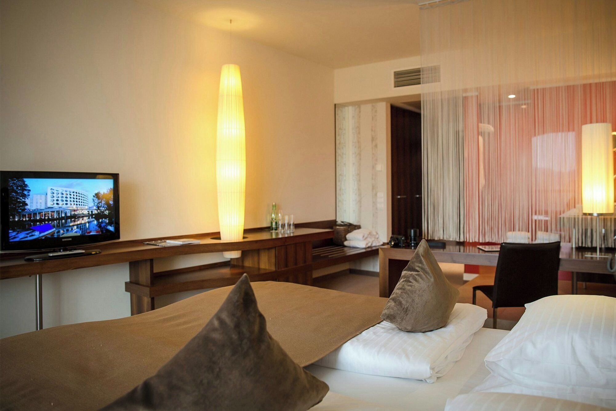 A hotel room with a bed, a desk with a television, and a large window with curtains.