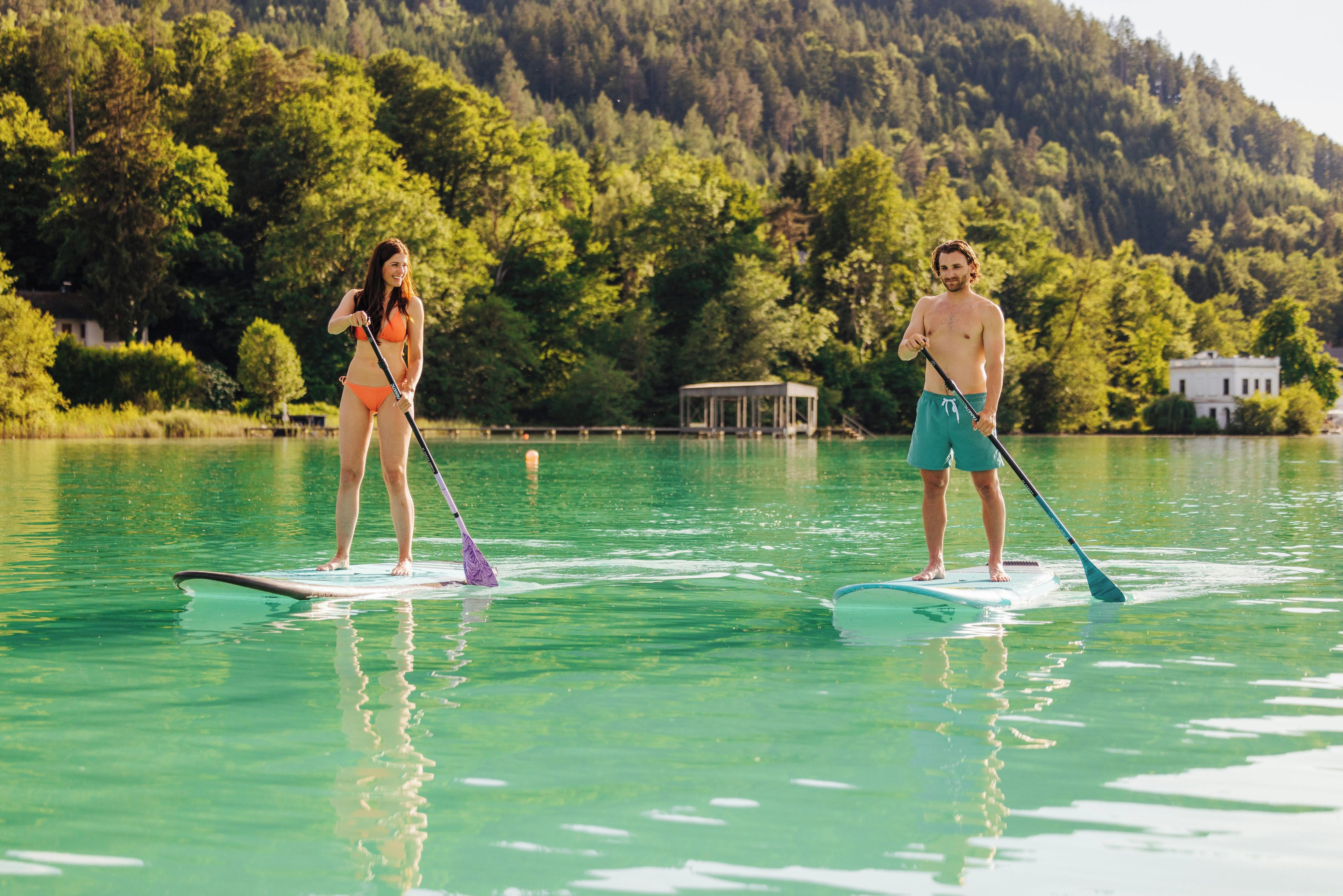 Two people paddleboarding on a calm lake with a forested hill in the background.