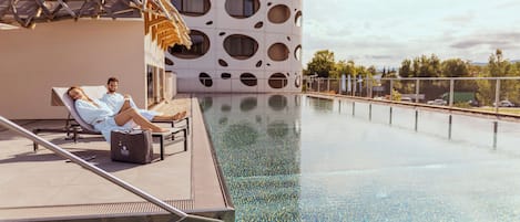 Een zwembadterras met twee ligbedden, een houten pergola en een modern gebouw met ronde ramen.