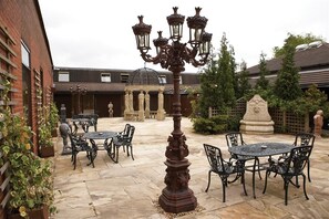 An outdoor patio with stone flooring, metal tables and chairs, a decorative iron lamp post, and a stone fountain. - Preston Leyland BW Signature Collection (Leyland)