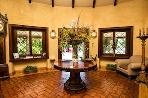 A room with a round wooden table, two windows with decorative frames, a chair, a clock, and a vase with flowers. - Hotel & Spa La Salve (Torrijos)