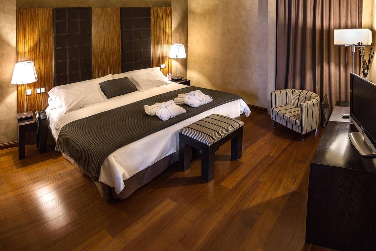 A hotel room with a large bed, two bedside lamps, a television, a bench, and a chair.