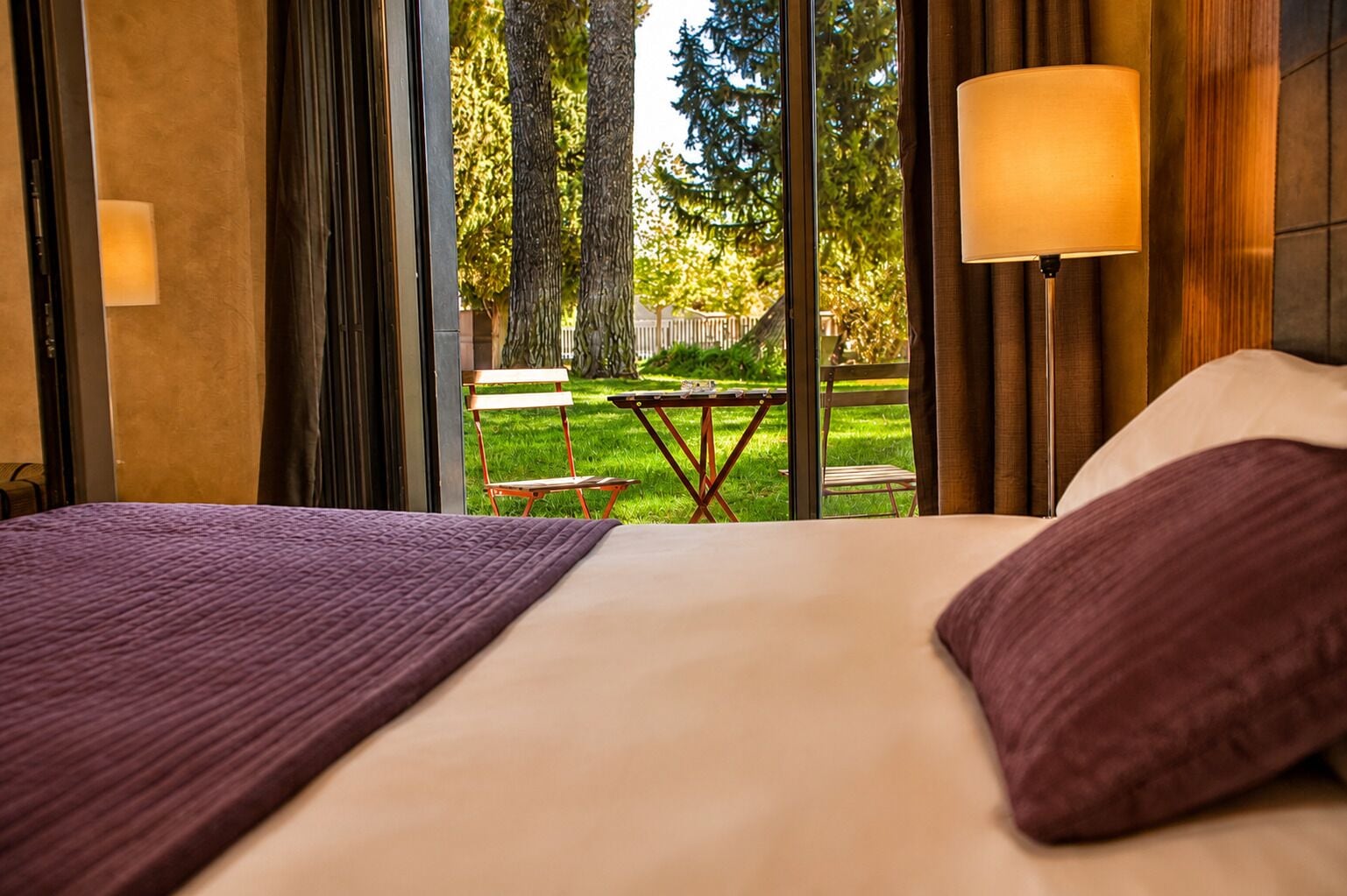 Quarto de hotel com uma cama, vista para um pátio com mesa e cadeiras, e um abajur na parede.