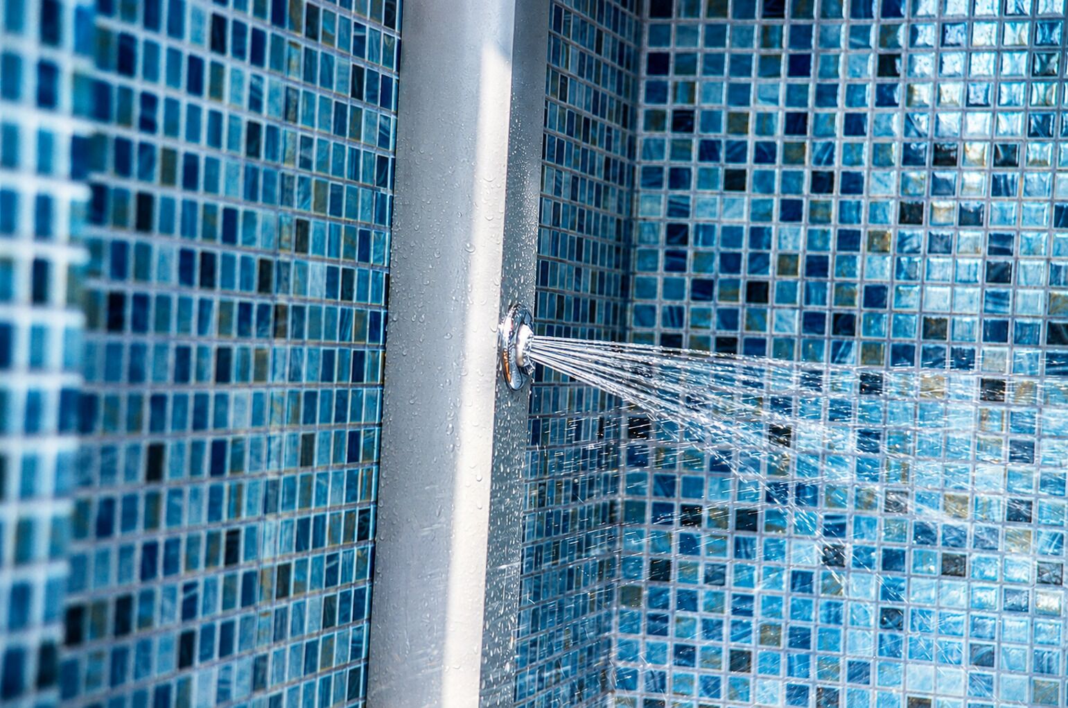 A shower with blue mosaic tiles and water flowing from a chrome fixture.