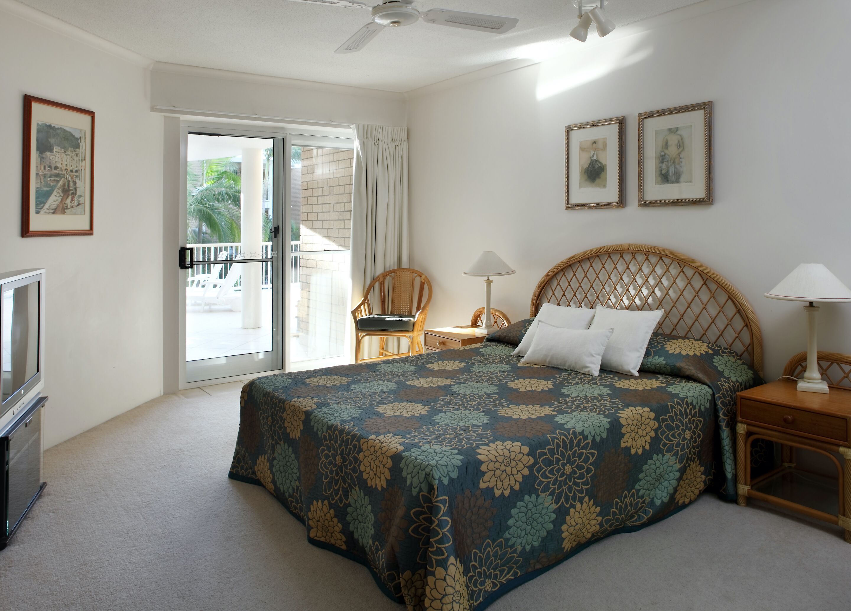 A hotel room with a large bed, a television, a balcony with a view of palm trees, and two framed pictures on the wall.