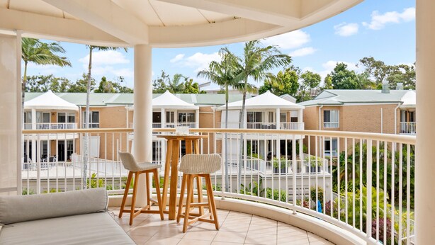 A balcony with a sofa and two chairs, overlooking a courtyard with palm trees and a building with a green roof. - Serenity Apartments Noosa (Noosa Heads)