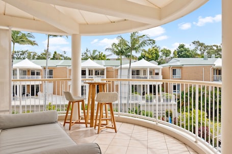 A balcony with a sofa and two chairs, overlooking a courtyard with palm trees and a building with a green roof.
