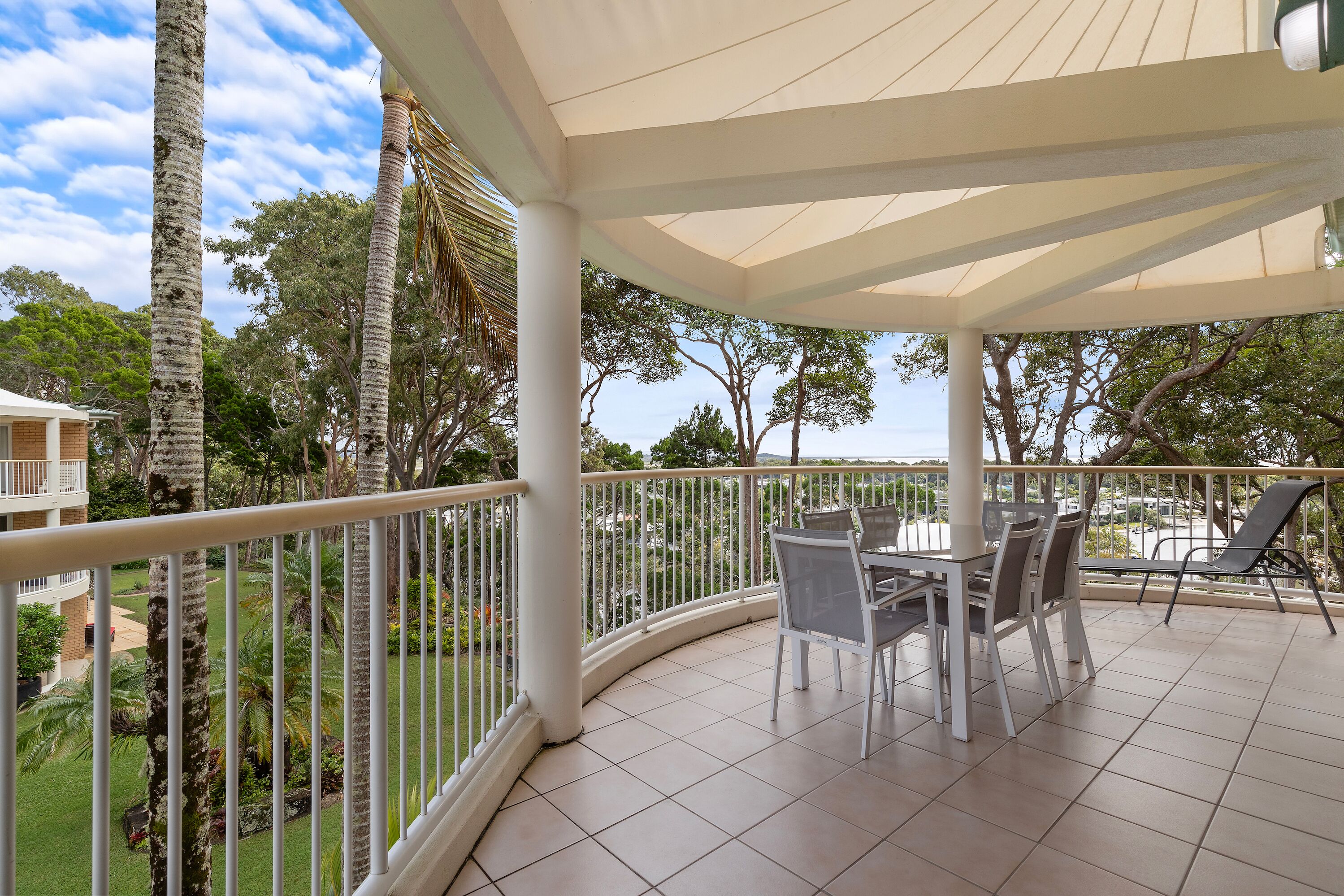 A covered outdoor area with a table and chairs, surrounded by a white railing, overlooking a landscaped garden and buildings.