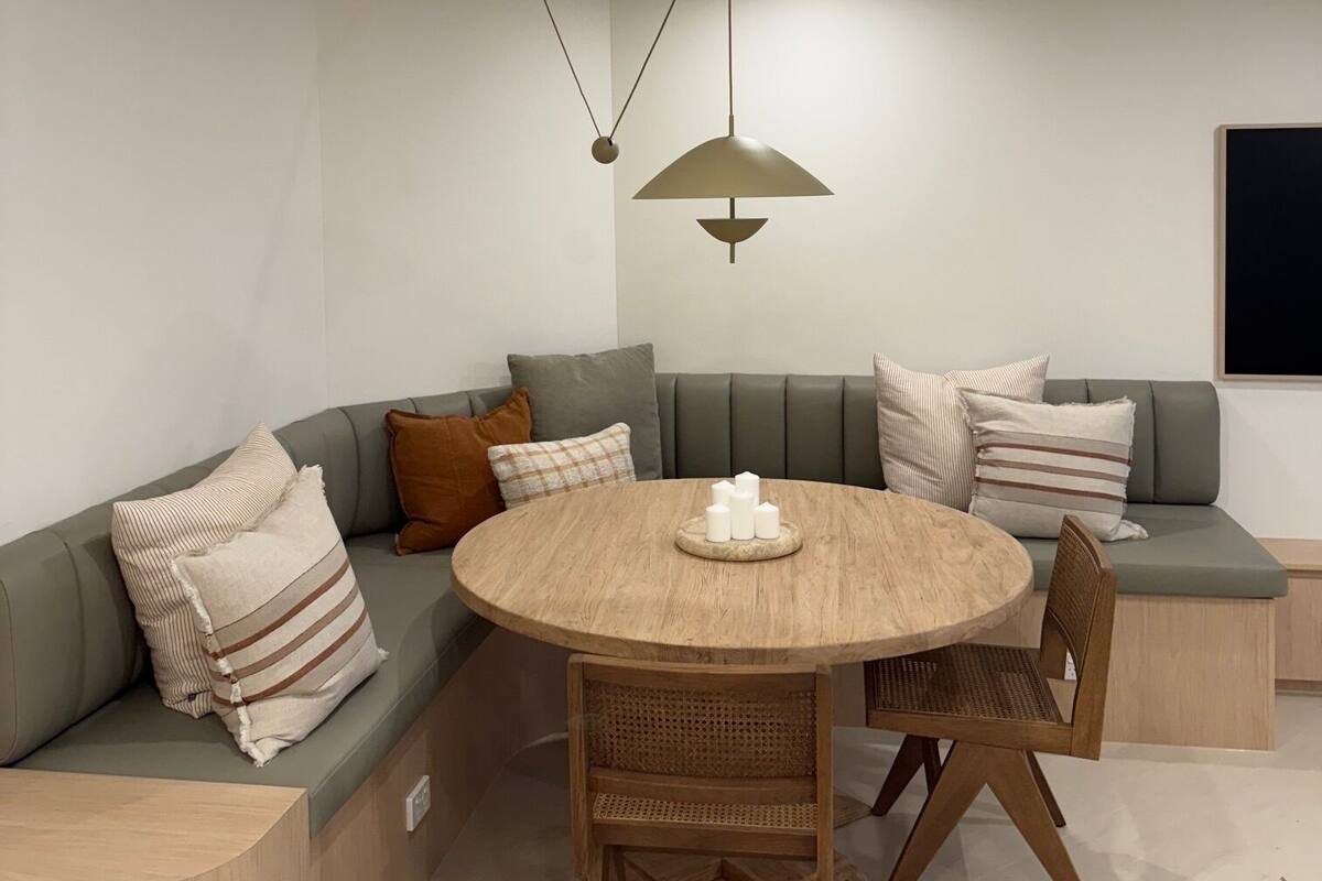 Ein modernes Wohnzimmer mit einem runden Holztisch, zwei Korbstühlen, einem grauen Ecksofa mit gestreiften Kissen und Pendelleuchten.