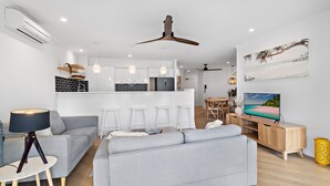 A modern living room with a grey sofa, a wooden TV stand, and a dining area with a table and chairs. - Serenity Apartments Noosa (Noosa Heads)