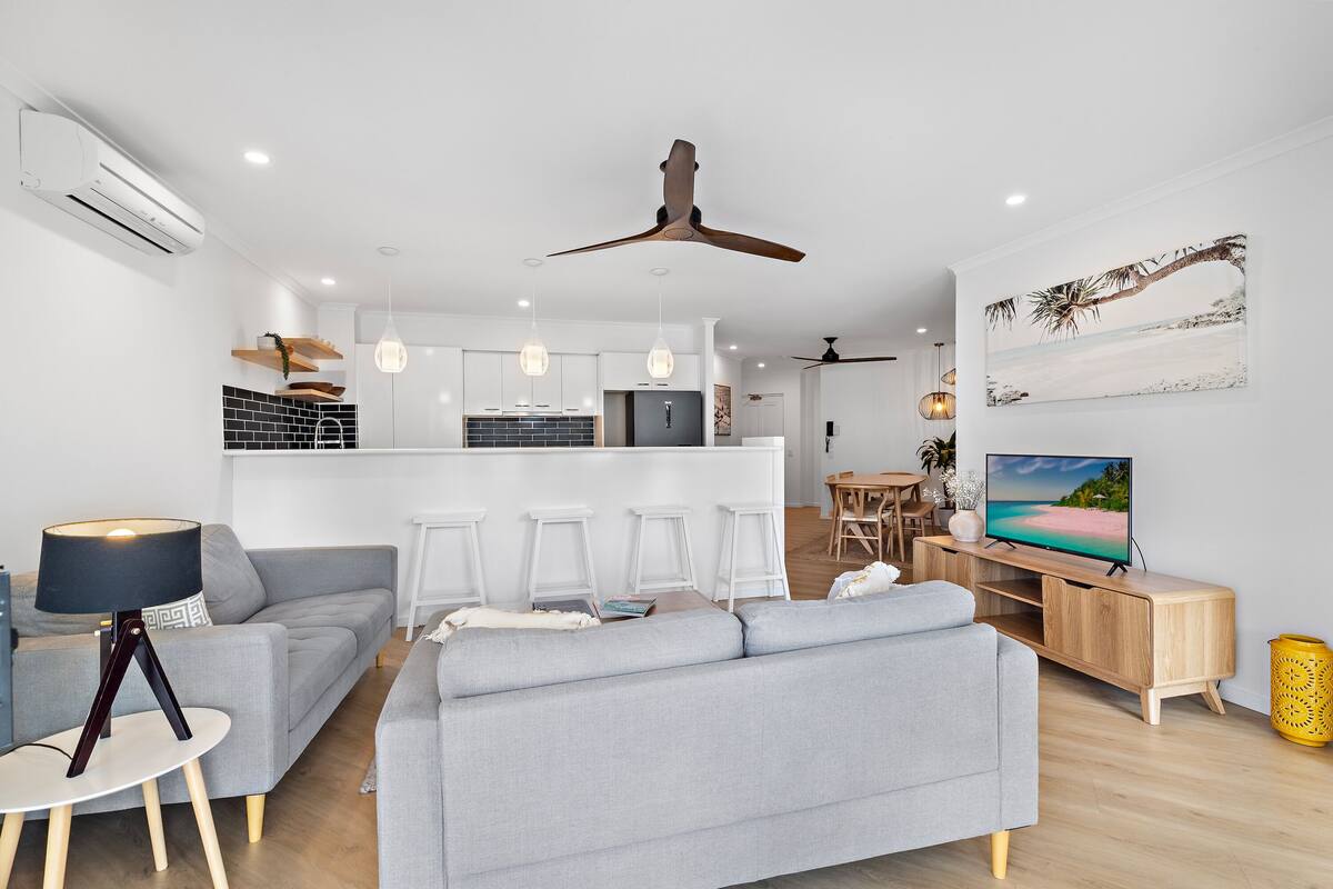Ein modernes Wohnzimmer mit grauem Sofa, einem Holz-TV-Schrank und einem Essbereich mit Tisch und Stühlen.