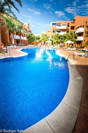 A swimming pool with clear blue water, surrounded by lounge chairs and palm trees, with buildings in the background.