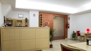 A reception area with a wooden reception desk, a plant, a fire extinguisher, and a closed door in the background. - Hotel Catania Town (Catania)