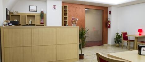 Un área de recepción con un mostrador de madera, una planta, un extintor y una puerta cerrada al fondo.