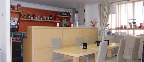 Uma área de refeições com mesa e cadeiras, uma cozinha com prateleiras cheias de garrafas e uma máquina de café.