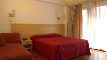 Una habitación de hotel con cabecera de madera, cama con colcha roja, mesita de noche con lámpara y ventana con cortinas.