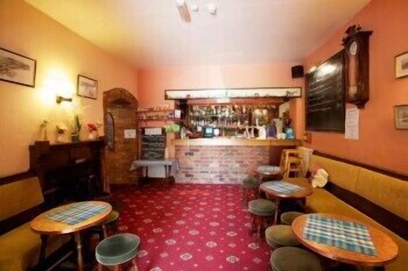 A cozy bar area with a red patterned carpet, wooden tables with checkered tablecloths, and a brick wall.