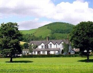 A white building with a gabled roof, surrounded by green fields and trees, with a hill in the background. - Abbey Grange Hotel (Llangollen)