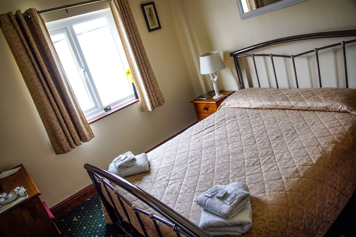 A neatly made bed with a quilted cover, a bedside lamp, and a window with curtains.