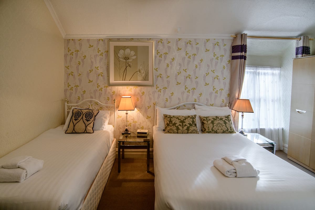 Quarto de hotel com duas camas, cada uma com travesseiro e toalha dobrada. Há duas mesas de cabeceira com abajures, um quadro emoldurado na parede e um guarda-roupa.