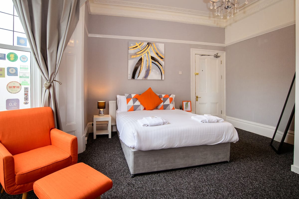 Quarto de hotel com uma cama, uma cadeira laranja, uma mesinha de centro com abajur e um quadro na parede.