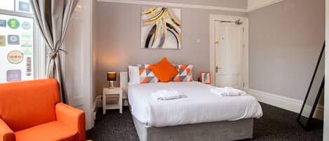 A hotel room with a bed, an orange chair, a small table with a lamp, and a painting on the wall.