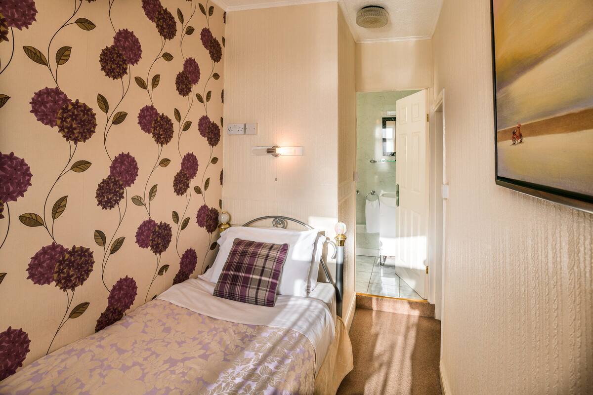 Quarto com cama, mesa de cabeceira, luminária na parede e porta para o banheiro.