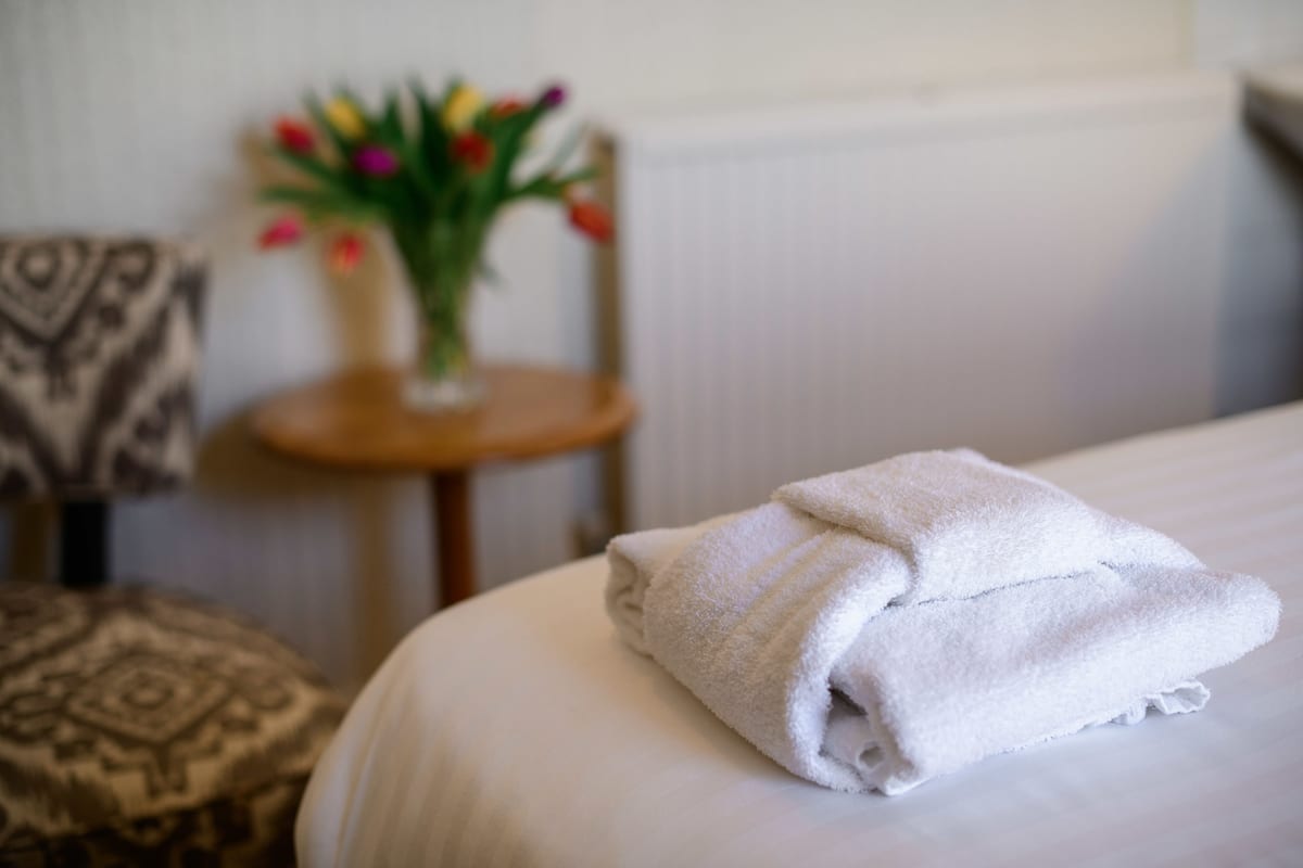 Cama bem arrumada com uma toalha branca dobrada, uma pequena mesa redonda com um vaso de flores e uma almofada estampada em uma cadeira.
