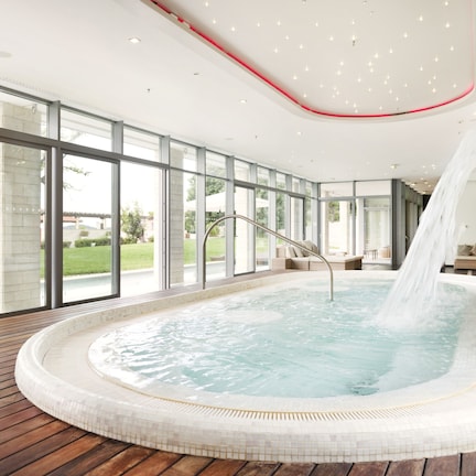 A modern indoor swimming pool with a wooden deck, large windows, and a waterfall feature.