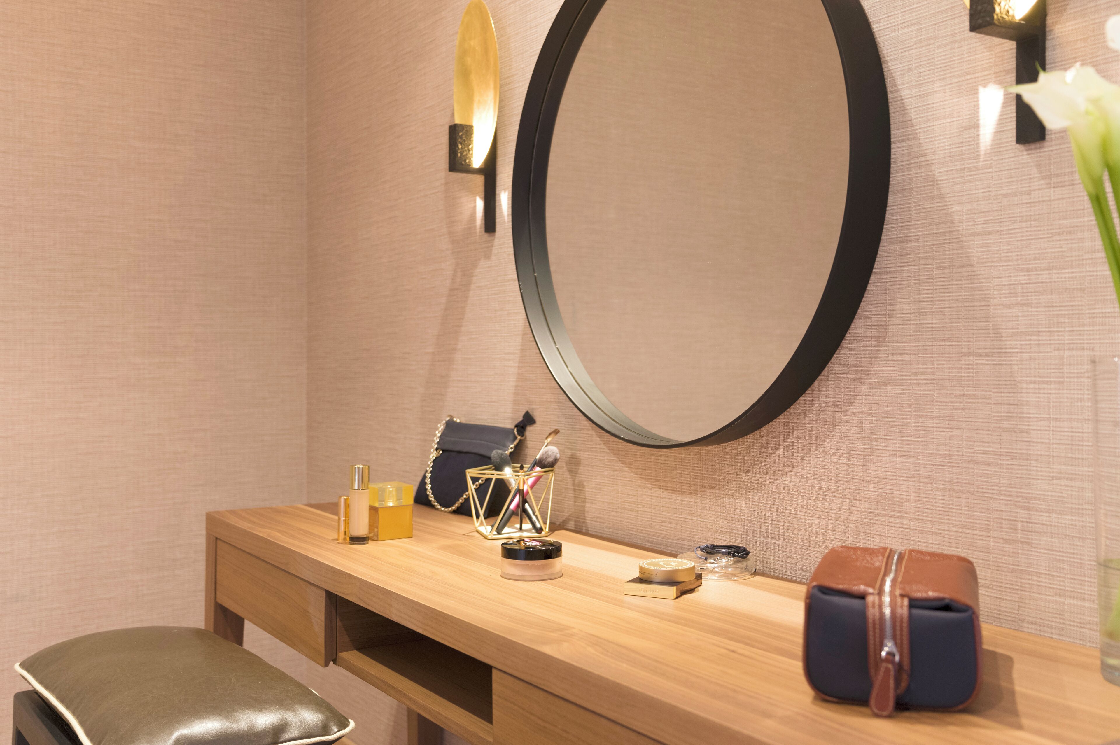 A wooden vanity with a round mirror, a bench, a small table with toiletries, and a handbag.