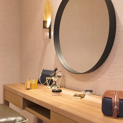 A wooden vanity with a round mirror, a bench, a small table with toiletries, and a handbag.