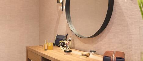 A wooden vanity with a round mirror, a bench, a small table with toiletries, and a handbag.