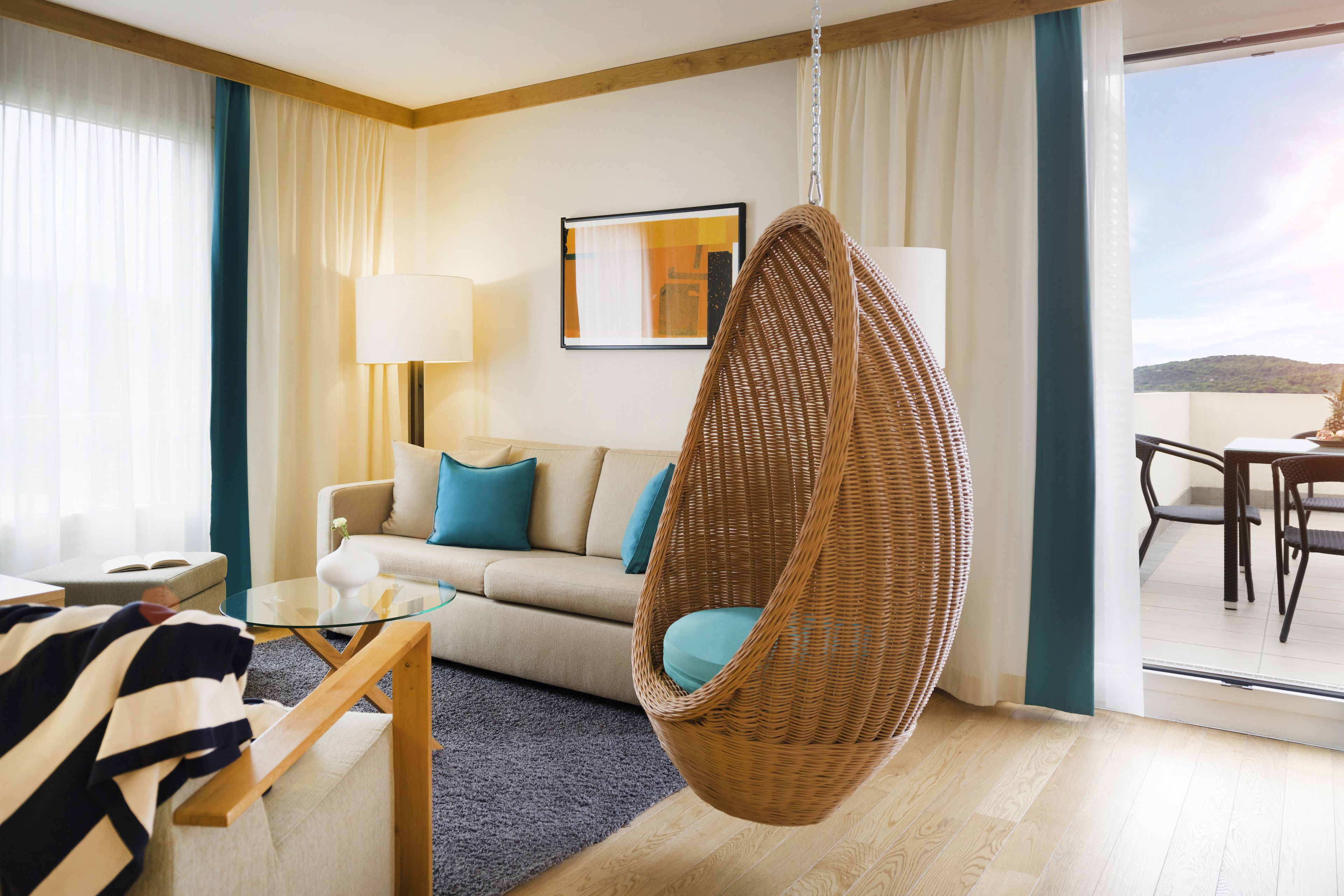Moderni hotellihuone, jossa on riipputuoli, sinisillä koristetyynyillä varustettu sohva, lasinen sohvapöytä ja näkymä ulos.