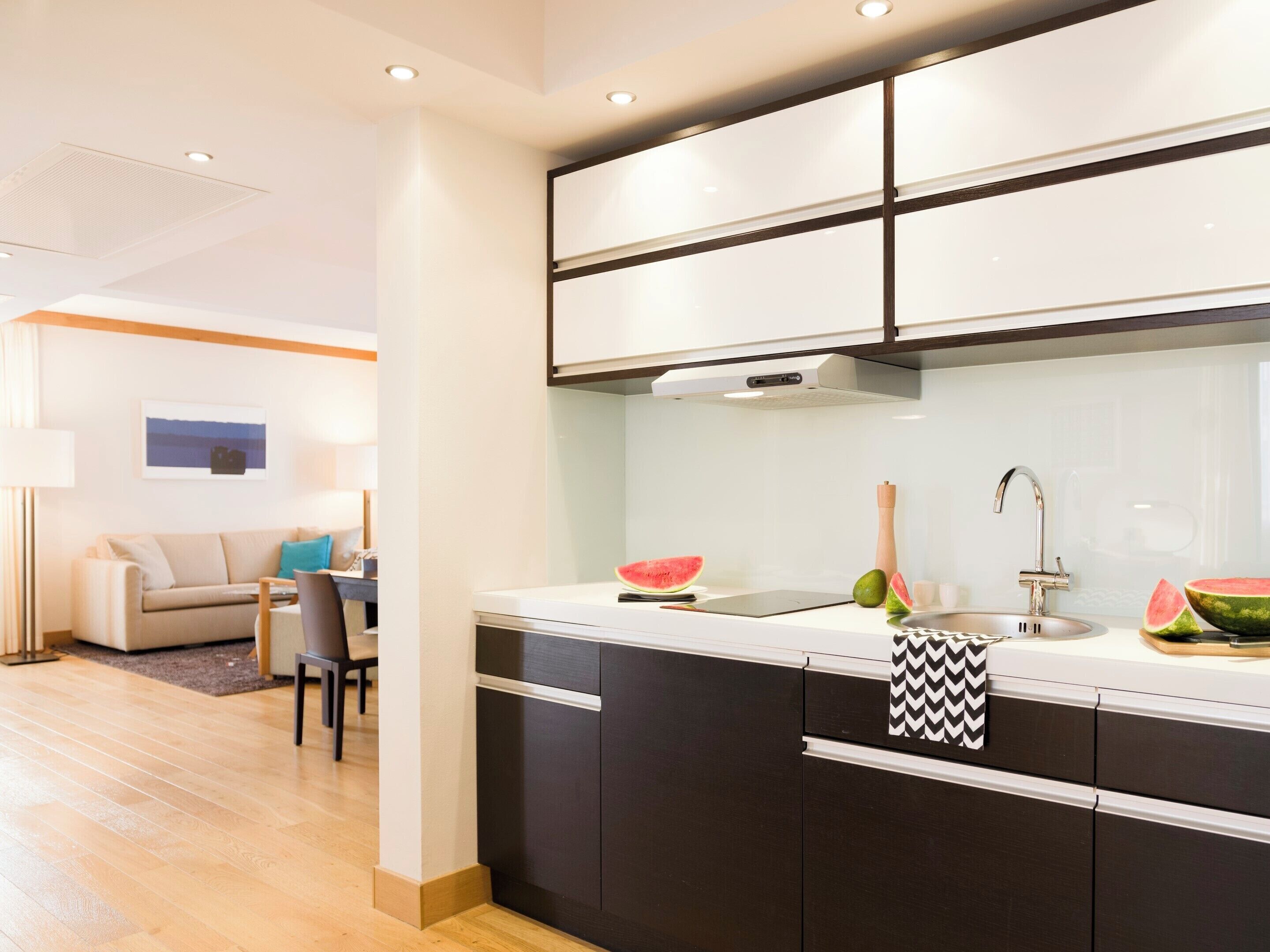 A modern kitchen with dark cabinets, a white countertop, and a stainless steel sink. There is a dining area with a sofa and a table in the background.