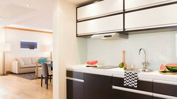 A modern kitchen with dark cabinets, a white countertop, and a stainless steel sink. There is a dining area with a sofa and a table in the background.