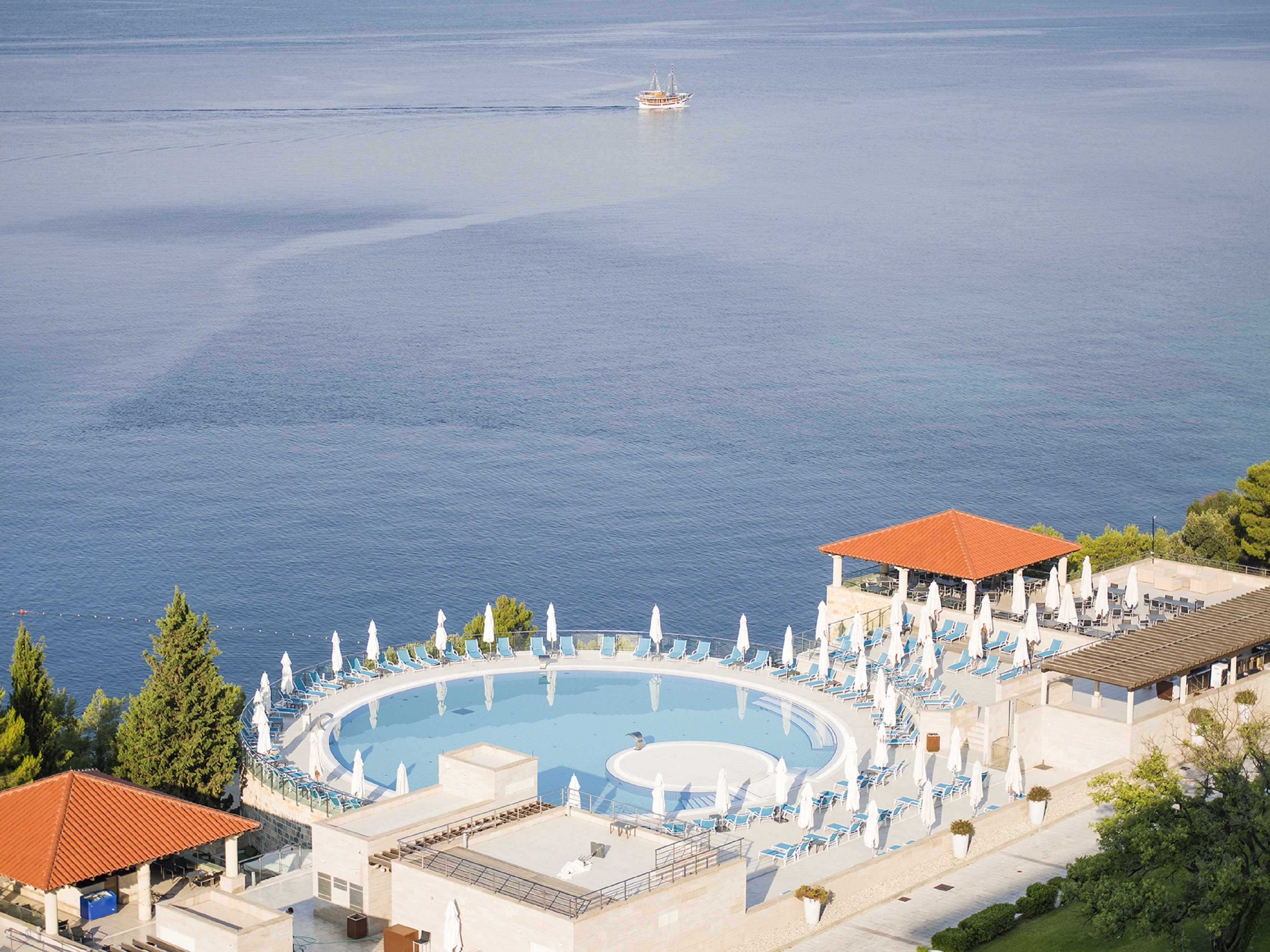 Un complex turístic amb una gran piscina circular envoltada de gandules i ombrel·les, amb vista a un mar tranquil.