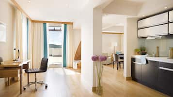 A modern hotel room with a wooden floor, a desk with a chair, a dining area with a table and chairs, and a kitchen with cabinets and a stove.
