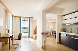 A modern hotel room with a wooden floor, a desk with a chair, a dining area with a table and chairs, and a kitchen with cabinets and a stove.